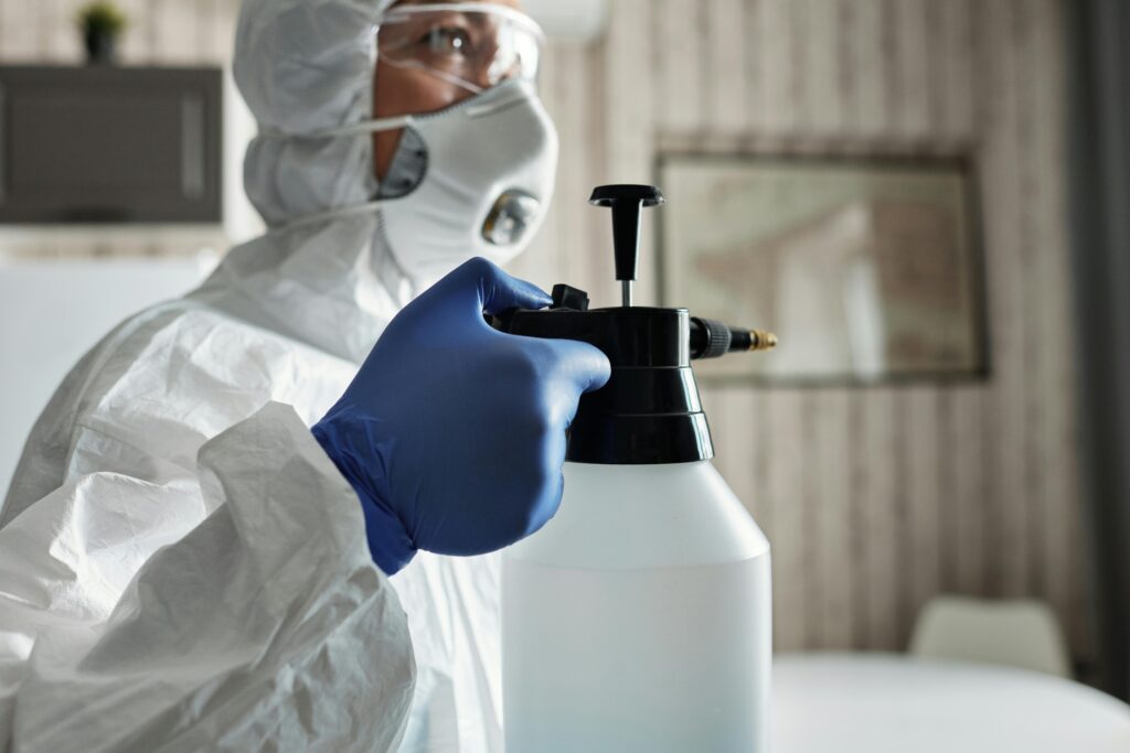 A person in protective gear with gloves and face mask using a spray bottle for disinfection indoors.